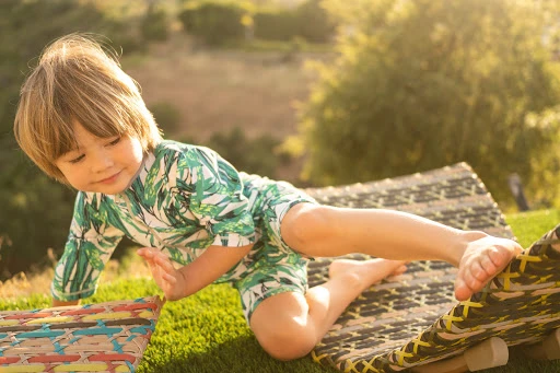 Blond child in tropical-print outfit plays on artificial grass with woven lounge chair in sunny outdoor setting. 