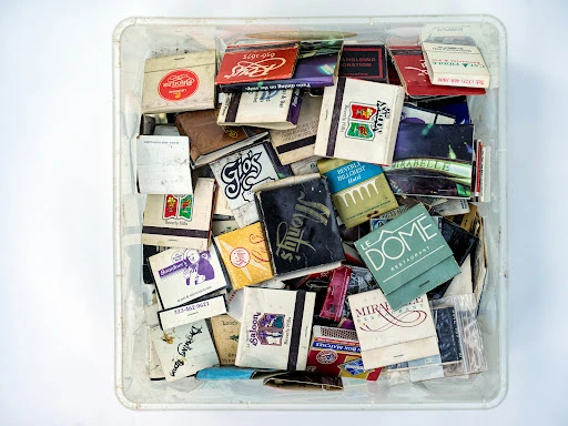 Collection of vintage matchbooks in a clear plastic container, featuring various designs and logos, set against a plain white background.