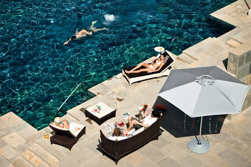 Overhead shot of people lounging and swimming in a turquoise pool near stone steps, with a neutral-toned patio umbrella providing shade.
