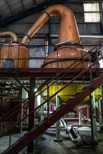 Interior view of a distillery with copper stills and metal stairs, showcasing industrial architecture and equipment for spirit production.