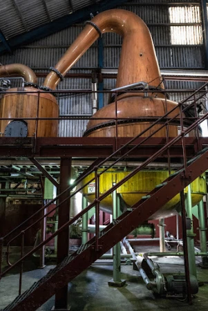 Interior view of a distillery with copper stills and metal stairs, showcasing industrial architecture and equipment for spirit production.
