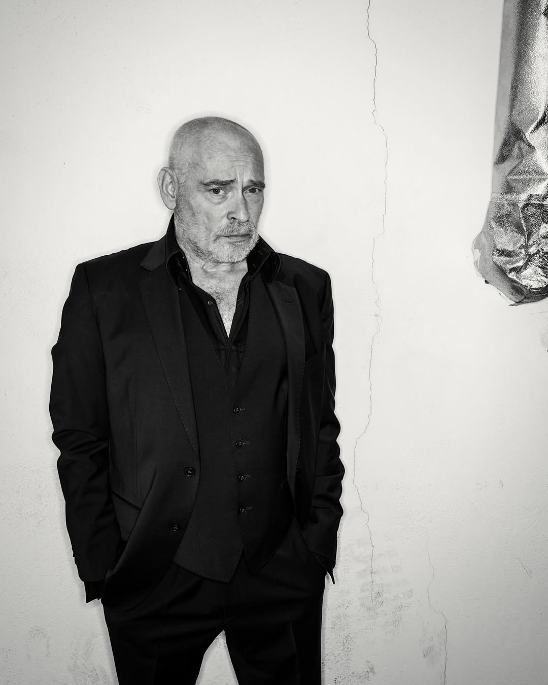 Black-and-white portrait of a mature man in a black suit and vest, looking serious against a white wall with subtle cracks.