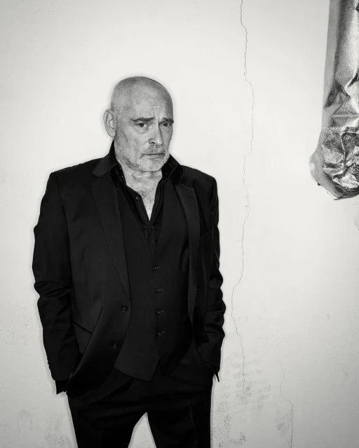 Black-and-white portrait of a mature man in a black suit and vest, looking serious against a white wall with subtle cracks.