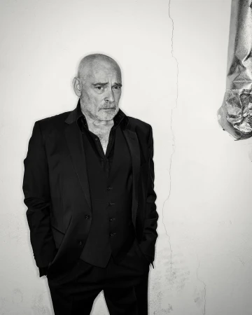 Black-and-white portrait of a mature man in a black suit and vest, looking serious against a white wall with subtle cracks.