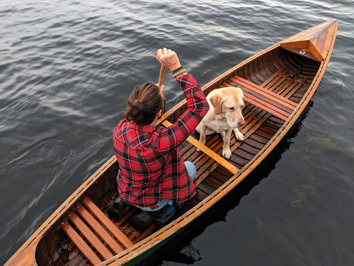 A man in a red plaid shirt paddles a wooden canoe with his dog sitting in the front, on a gray lake, taken from an overhead perspective.