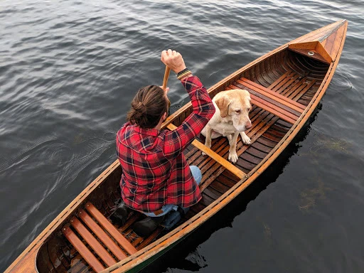 A man in a red plaid shirt paddles a wooden canoe with his dog sitting in the front, on a gray lake, taken from an overhead perspective.