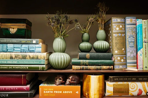 Stacked hardcover books in various colors on a brown shelf, interspersed with green vases, skulls, and a gold container.