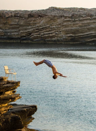 Man dives into the ocean from a rocky cliff next to a white chair, with a layered rock formation in the background under a soft sky.