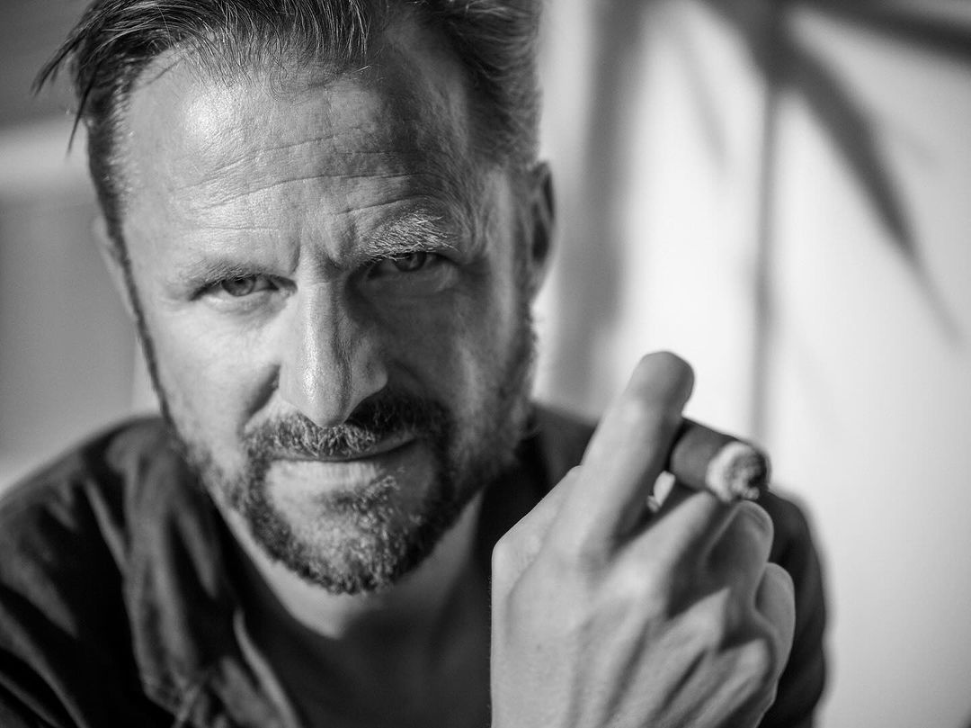 Black and white portrait of a middle-aged man holding a lit cigar indoors, close-up shot with soft lighting and a neutral expression.