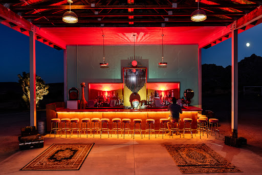 Outdoor bar at night with a lone patron sitting at the counter, illuminated by red lighting, casting a warm glow on the stools.