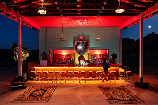 Outdoor bar at night with a lone patron sitting at the counter, illuminated by red lighting, casting a warm glow on the stools.