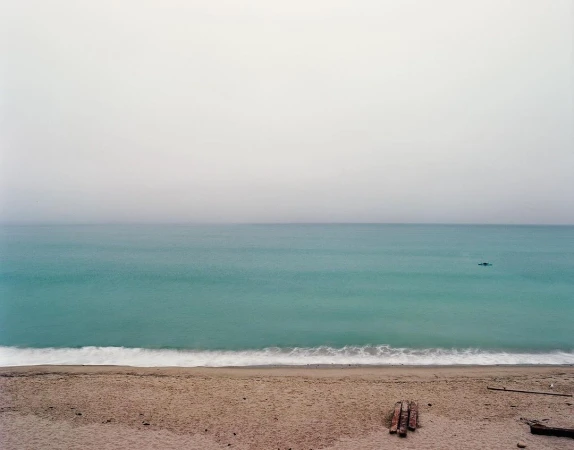 A wide coastal landscape under an overcast sky featuring a sandy beach, gentle teal ocean waves, and a calm horizon.