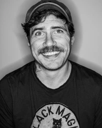 Black and white portrait of a smiling man with a mustache wearing a baseball cap and a graphic t-shirt.