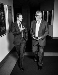 Two men in suits walking down an office hallway, talking and smiling in a monochrome portrait by Rainer Hosch.