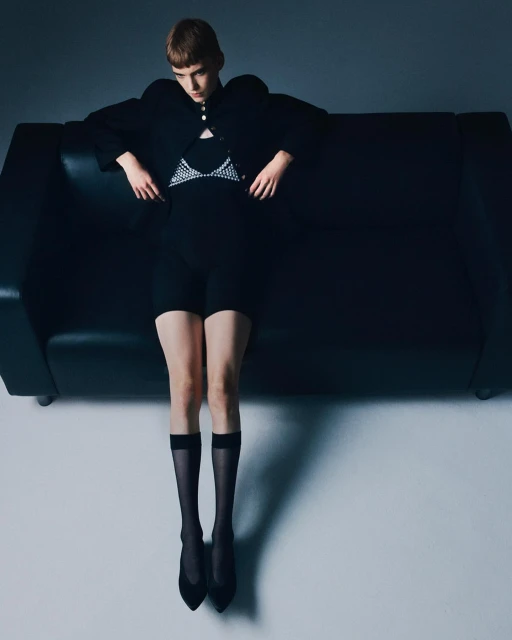 A slender woman with short brown hair, wearing a black top, shorts, jacket, sheer socks, and heels, poses on a black couch in a studio.