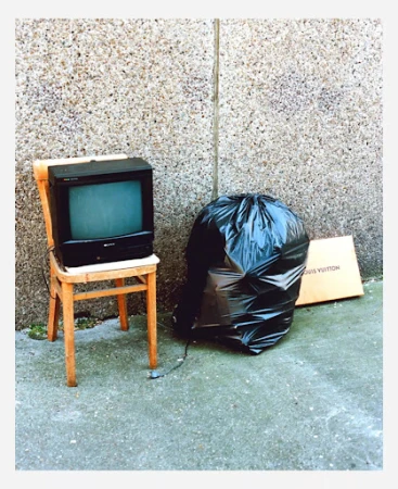 An old CRT television rests on a wooden chair next to a black garbage bag and a cardboard box against a textured concrete wall.