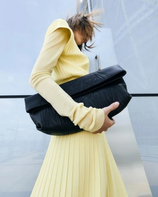 Woman in yellow dress holds a large black leather clutch with textured details in front of a silver metal facade.