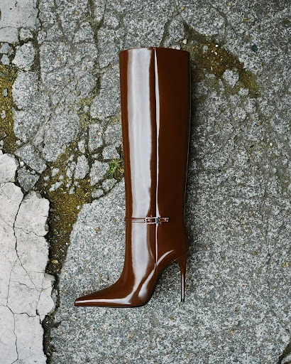 Brown, high-heeled, knee-high boot with a silver buckle on a textured gray concrete surface with some discoloration and small green patches.