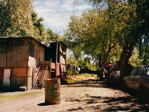 A weathered wooden shack with a rusty barrel in a rural, overgrown environment, featuring a vintage car and clothesline in the background.