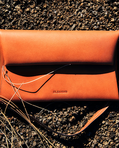 An orange leather Jil Sander clutch bag, accented with thin strings, rests on a textured, dark brown pebbled surface outdoors.