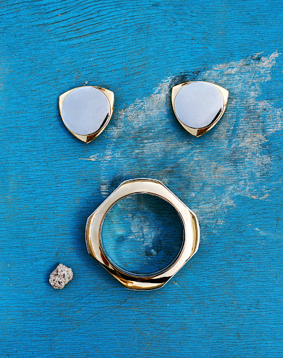 Overhead shot of a golden bracelet, two shield-shaped objects, and a grey rock on a distressed blue wooden surface. Product photography.