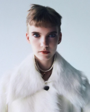 Portrait of a model wearing a white faux fur coat and layered pearl necklaces against a light studio backdrop