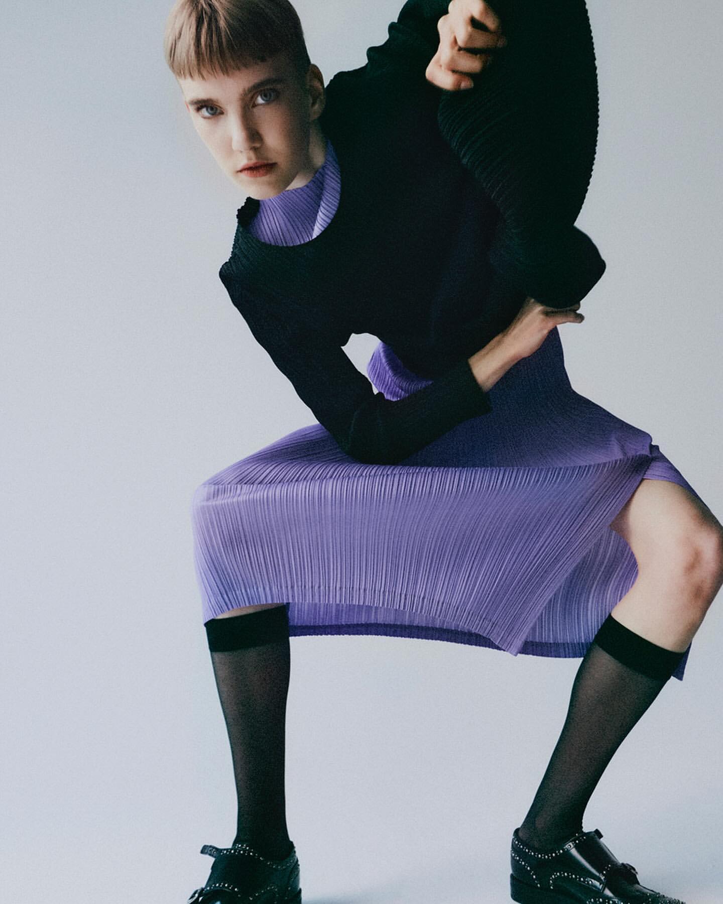 Fashion shot of an adult female with a severe expression. The subject wears a black top, purple skirt and black socks with black loafers.