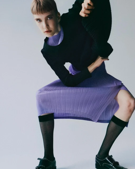 Fashion shot of an adult female with a severe expression. The subject wears a black top, purple skirt and black socks with black loafers.
