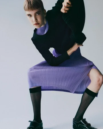 Fashion shot of an adult female with a severe expression. The subject wears a black top, purple skirt and black socks with black loafers.