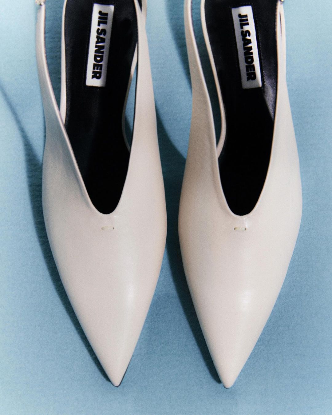 Close-up product shot of two white leather Jil Sander heeled pumps, with pointed toe and slingback, against a light blue background.