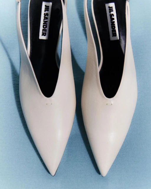Close-up product shot of two white leather Jil Sander heeled pumps, with pointed toe and slingback, against a light blue background.
