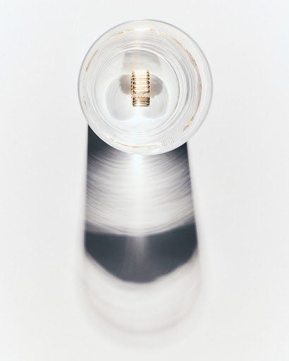 A clear glass jar with a metal component inside stands on a white surface, casting a blurry shadow in a minimalist still life.