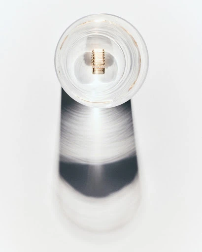 A clear glass jar with a metal component inside stands on a white surface, casting a blurry shadow in a minimalist still life.