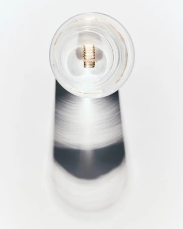 A clear glass jar with a metal component inside stands on a white surface, casting a blurry shadow in a minimalist still life.