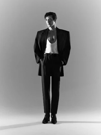 A man in an oversized black blazer and trousers stands against a grey seamless studio backdrop.