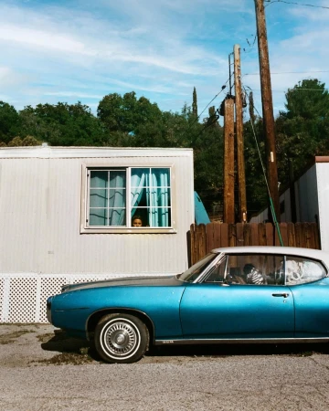 Vintage turquoise car parked near a mobile home with a person looking out the window under a sunny blue sky with scattered clouds.