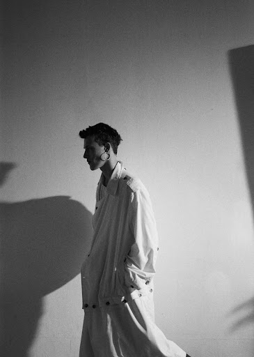 Monochrome profile of a person wearing an oversized white coat and earring, walking in front of a white wall with subtle shadows.