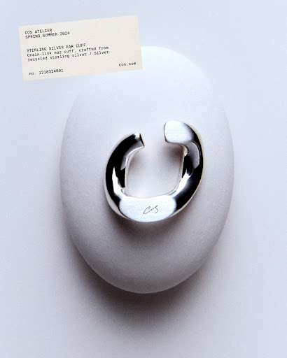 Shiny silver ear cuff displayed on a smooth, light-grey stone against a gradient grey backdrop, accompanied by a product information card.