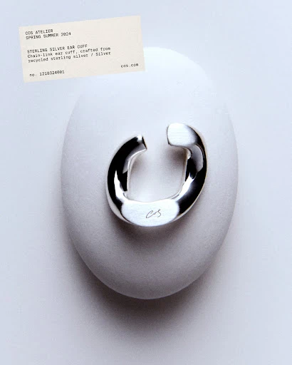Shiny silver ear cuff displayed on a smooth, light-grey stone against a gradient grey backdrop, accompanied by a product information card.