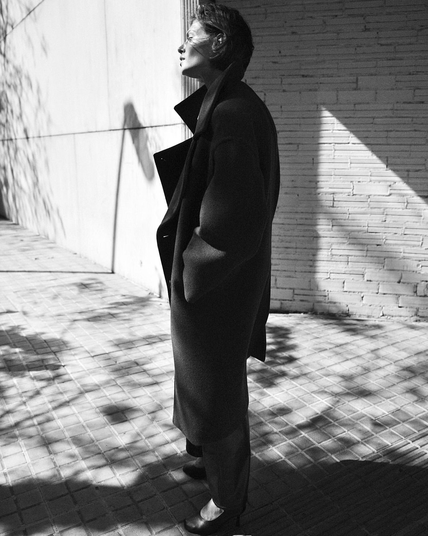 Black and white outdoor portrait of a woman wearing a long coat, standing on a stone surface next to a textured wall with tree shadows.