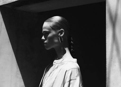 Monochrome portrait of a woman with slicked-back hair and geometric earrings, standing in stark sunlight against a dark background.