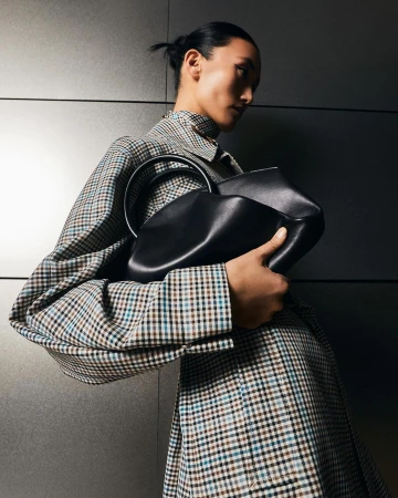 Adult female model presents a black leather handbag with a metal handle, wearing a checkered coat in a dimly lit studio setting.