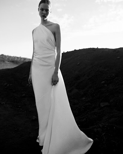 Model in a white one-shoulder gown standing outdoors on a dark terrain under an overcast sky.