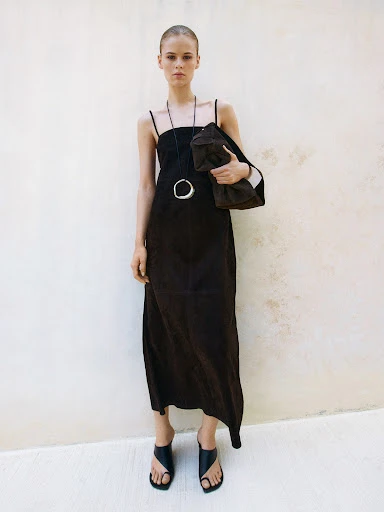 A fair-skinned woman poses in a full-length, dark brown dress and black sandals, holding a bag against a light wall indoors.