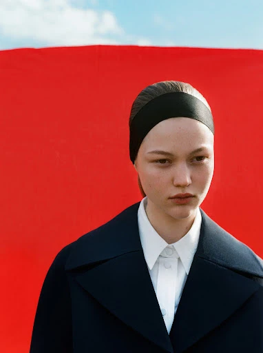 Close-up of a young fair-skinned adult woman with a black headband, wearing a navy blue coat, posing against a red and blue sky background.