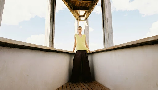 Model standing on a wooden balcony wearing a yellow sleeveless top and dark trousers captured from a low angle.
