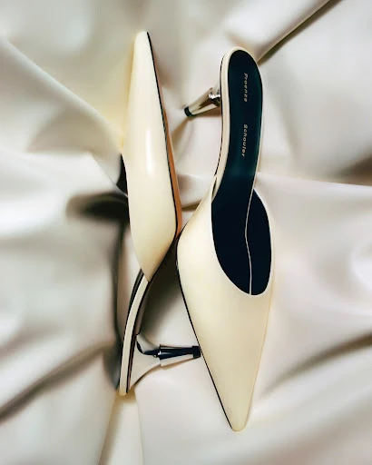 Cream-colored pointed-toe mule heels by Proenza Schouler, with small metallic heels, elegantly placed on a draped silk fabric backdrop.