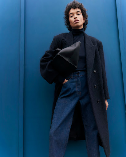 A fashion portrait of a woman wearing a black coat, blue jeans, and a black top, holding a black tote bag against a blue wall.