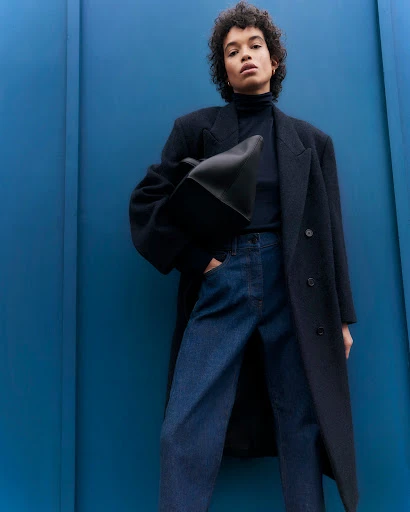 A fashion portrait of a woman wearing a black coat, blue jeans, and a black top, holding a black tote bag against a blue wall.
