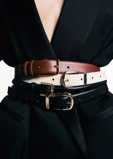 Three leather belts in shades of brown, cream, and black with gold buckles are stacked around a person's waist, paired with a black blazer.